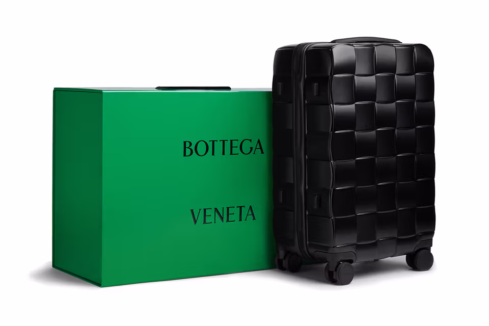 A black carry-on suitcase with a woven design, placed next to a green box featuring the Bottega Veneta logo.